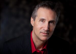A headshot photo of George Mitchell, a man with gray hair, a red button-down shirt, and a black suit jacket, smiling at the camera.