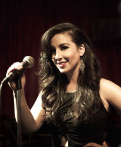 A portrait of Gina Saputo, a woman with long, dark, wavy hair, wearing a sleeveless black top and dangling earrings, smiling while holding a microphone on a stand.
